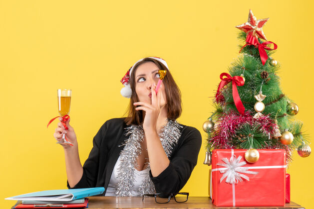 圣诞老人情绪化快乐迷人的女士穿着西装 戴着圣诞老人的帽子和新年的装饰品 在黄色的办公室里举杯美丽葡萄酒成人