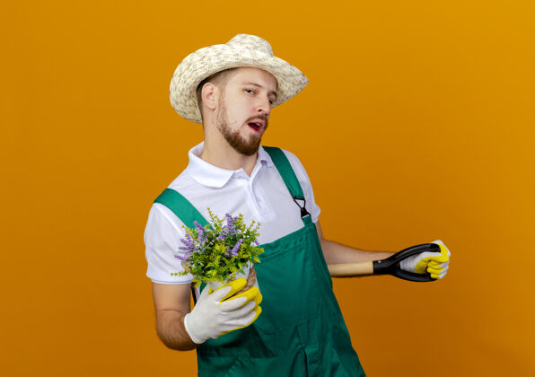 制服自信的年轻英俊的斯拉夫园丁穿着制服戴着帽子和园艺手套手持铁锹和花盆看起来与世隔绝花盆穿花园
