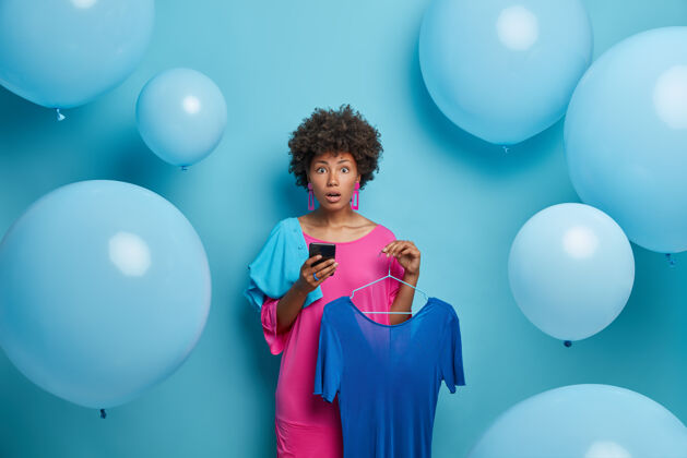 成人惊讶的黑皮肤女性选择服装 把蓝色的衣服挂在衣架上 使用智能手机 在时尚精品店网上购物 准备约会或派对 隔离在蓝色的墙上衣服鞋子惊喜