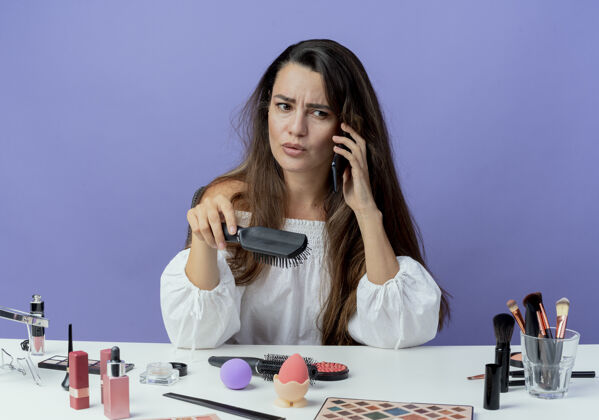 漂亮恼怒的美女拿着化妆工具坐在桌边拿着梳子讲电话看着紫色墙上孤立的一面电话烦恼坐着