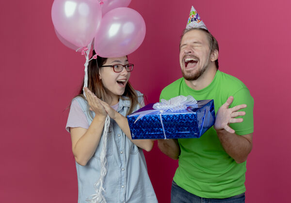 帽子快乐的年轻女孩拿着氦气球看着兴奋的年轻人戴着派对帽拿着礼品盒隔离在粉红色的墙上生日氦年轻