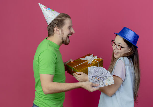 请戴派对帽的欢笑青年给戴蓝色派对帽的快乐青年送钱 把礼盒隔离在粉红色的墙上粉色年轻蓝色