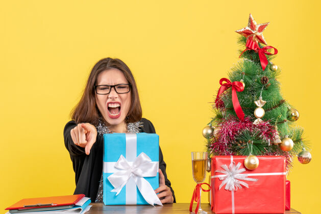 人情绪混乱的商务女士穿着西装 戴着眼镜 拿着礼物对着别人大喊大叫 坐在一张桌子旁 桌子上放着一棵圣诞树喊叫快乐礼物