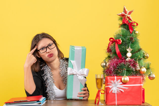 女士困惑的商务女士穿着西装 戴着眼镜展示她的礼物 坐在一张桌子旁 桌子上放着一棵圣诞树漂亮花束人
