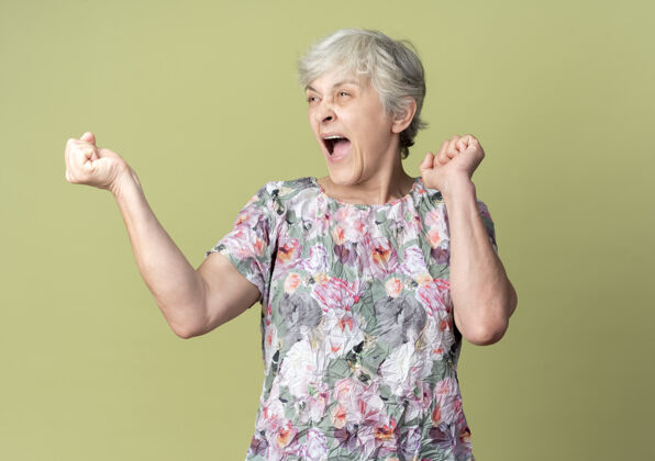 老年人快乐的老妇人举起拳头看着隔离在橄榄绿墙上的一面举起来橄榄色女人