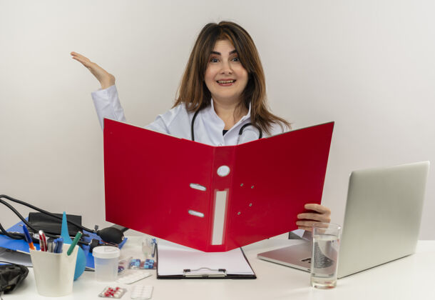 女人快乐的中年女医生穿着医用长袍和听诊器坐在办公桌旁 拿着医疗工具剪贴板和笔记本电脑夹 显示空手孤立手桌子工具