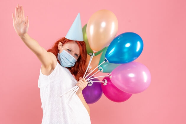 年正面图：年轻女性手持彩色气球 戴着无菌面具站在粉红色的椅子上举行庆祝生日