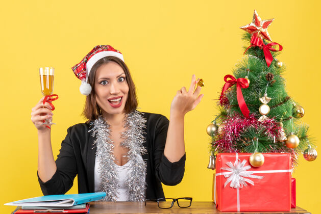 成人情绪化的快乐满足的迷人女士穿着西装 戴着圣诞老人的帽子和新年的装饰品 在黄色的办公室里举杯庆祝圣诞老人西装年