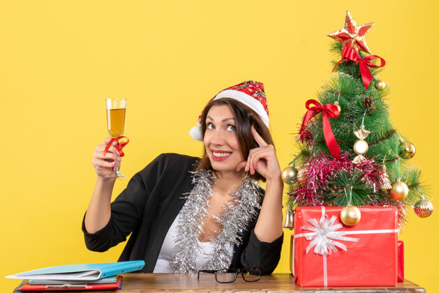 快乐情绪迷人的女士穿着西装 戴着圣诞老人的帽子和新年的装饰品 在黄色的办公室里举杯庆祝酒帽子成人