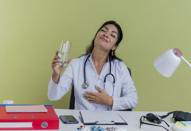 长袍高兴的年轻女医生穿着医用长袍和听诊器坐在办公桌旁 手放在胸前 抱着一杯水 闭着眼睛 与世隔绝工具医生胸部