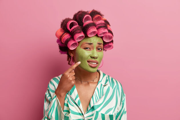 治疗不高兴的少数民族妇女的照片指向脸上的问题区域 指在脸颊和显示丘疹 戴绿色保湿面膜 应用发夹 穿休闲睡衣美丽肤色长袍年轻