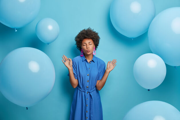 冷时尚迷人的女人举手 嘴唇圆润 心情愉快 拿着气球对着蓝色的墙壁摆姿势 在装饰好的房间里拍照 穿着漂亮的长裙明亮手掌衣服