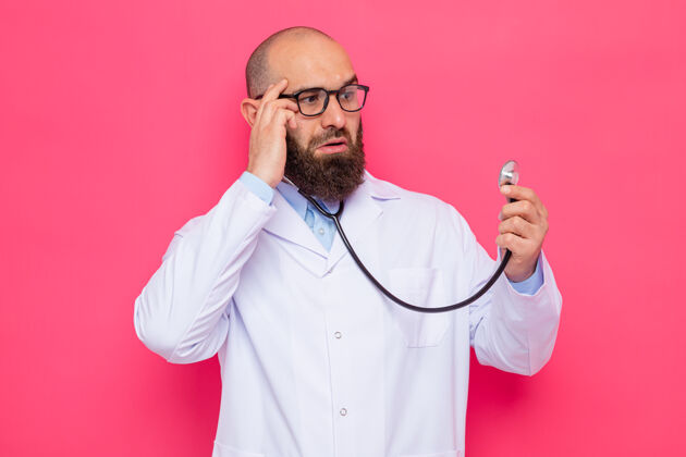 胡子一位留着胡子的医生 穿着白大褂 戴着眼镜 手持听诊器 站在粉色的背景上惊奇地看着它眼镜听诊器支架