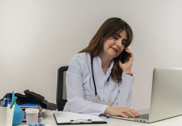 羽绒服高兴的中年女医生穿着医用长袍和听诊器坐在办公桌旁 拿着医疗工具剪贴板和笔记本电脑讲电话 用笔记本电脑隔着头往下看医疗成人女士
