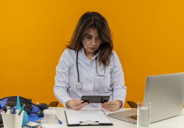 坐着集中的中年女医生穿着医用长袍和听诊器坐在办公桌旁 拿着医疗工具 剪贴板和笔记本电脑 孤立地看着手机手机成人女士