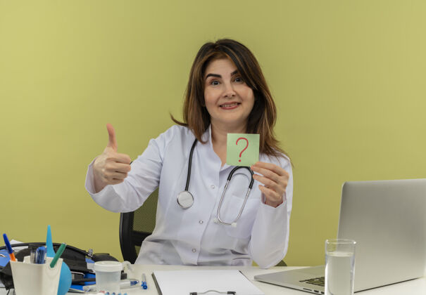 抱着微笑的中年女医生穿着医用长袍和听诊器坐在办公桌旁 手里拿着医疗工具和笔记本电脑 手里拿着问号 孤立地竖起大拇指微笑笔记本电脑医生