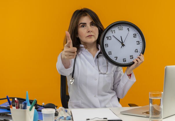 指点自信的中年女医生穿着医用长袍和听诊器坐在办公桌旁 带着医疗工具剪贴板和笔记本电脑 把时钟的指针隔离开来桌子抱着女士