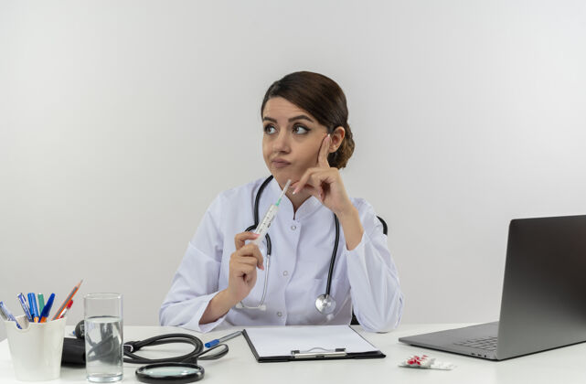 人体贴的年轻女医生穿着医用长袍和听诊器坐在办公桌旁 拿着医疗工具和手提电脑 拿着注射器 一边看着一边把手指贴在脸上坐着保持医疗