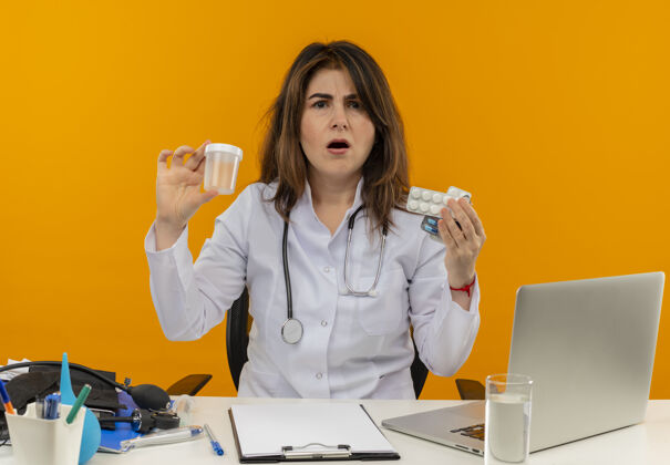 医疗令人印象深刻的中年女医生穿着医用长袍 听诊器坐在办公桌旁 带着医疗工具剪贴板和笔记本电脑 手里拿着隔离的药品和烧杯中年女士坐着