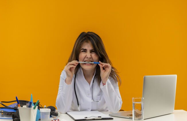 女性恼怒的中年女医生穿着医用长袍和听诊器坐在办公桌前 手里拿着医疗工具剪贴板和笔记本电脑咬笔 看上去与世隔绝医生听诊器笔记本电脑
