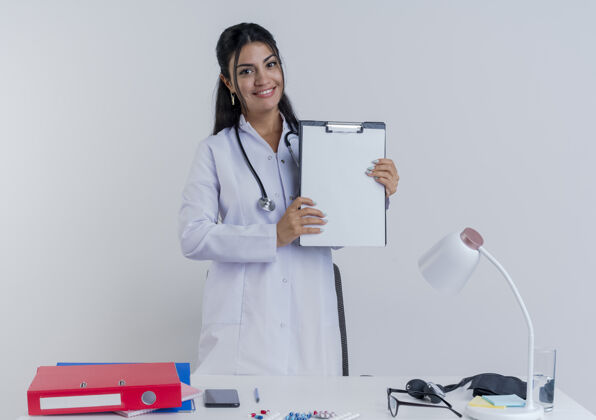 成人面带微笑的年轻女医生穿着医用长袍和听诊器坐在办公桌旁 手里拿着医疗工具 看上去与世隔绝长袍桌子年轻人