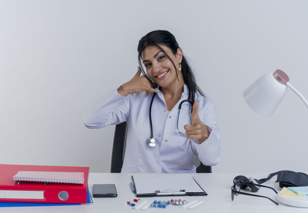 电话面带微笑的年轻女医生穿着医用长袍和听诊器坐在办公桌旁 手里拿着医疗工具 做着呼叫手势和手势坐着桌子长袍