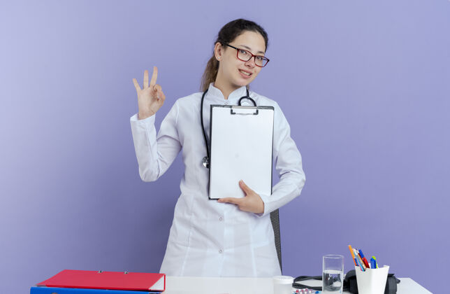 听诊器面带微笑的年轻女医生 穿着医用长袍 戴着听诊器和眼镜 站在桌子后面 拿着医疗工具 拿着剪贴板 看起来很孤立微笑穿紫色
