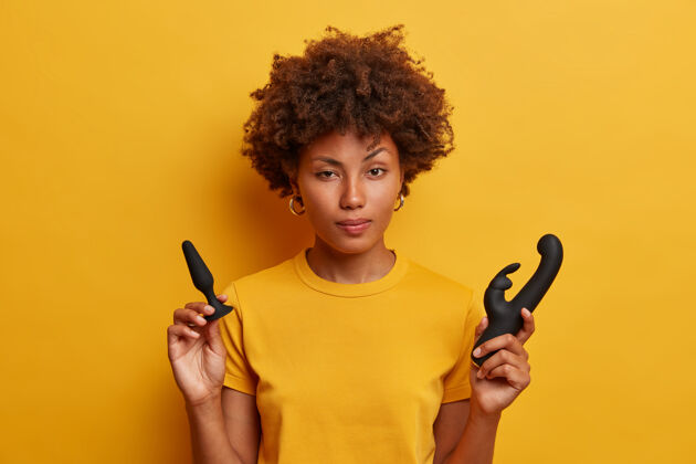 休闲犹豫不决的非洲裔美国女性在插入式游戏前拿着屁股塞热身 兔子形状的振动器刺激阴道 穿着黄色t恤室内有性玩具的年轻女性臀部室内插头