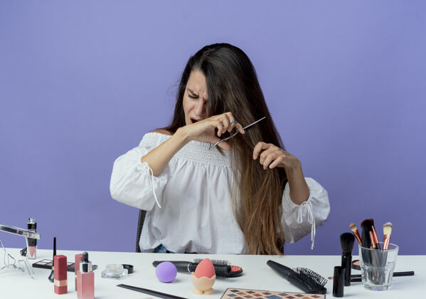 剪恼怒的美女拿着化妆工具坐在桌边假装用剪刀剪头发孤立在紫色的墙上坐化妆剪刀