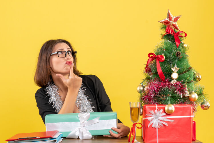 套装一位穿着西装 戴着眼镜的商务女士惊讶地拿着礼物 坐在办公室里一张桌子旁 桌子上放着一棵圣诞树惊喜笔记本电脑微笑