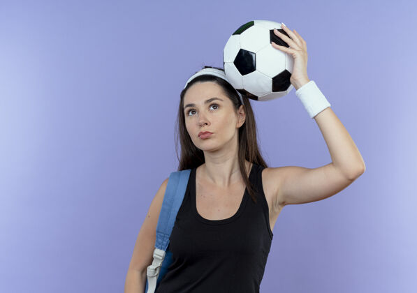 头戴着头巾 背着背包 头上举着足球的年轻健身女士站在蓝色背景下 看起来很自信头带抱球