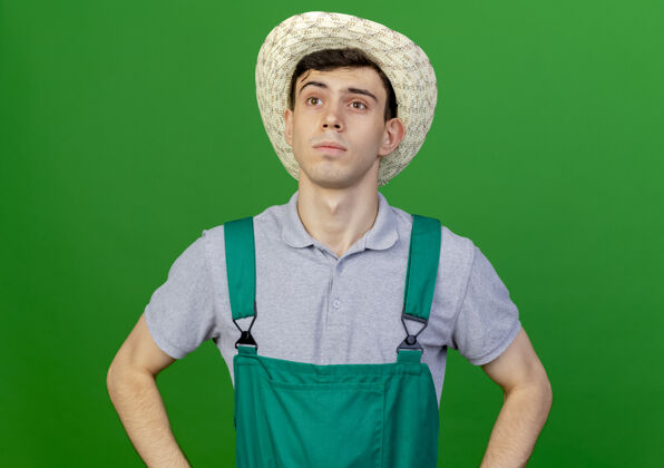 空间自信的年轻男园丁戴着园艺帽 双手放在腰上 看着绿色背景上与世隔绝的一面 留着空间年轻绿色园艺