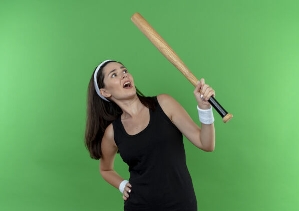 困惑戴着头巾拿着棒球棒站在绿色背景下困惑地看着它的年轻健身女士站健身头带