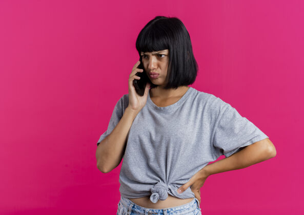 复制恼怒的年轻黑发白人女孩把手放在腰上讲电话 看着粉红色背景上孤立的一面和复印空间电话粉红色壁板