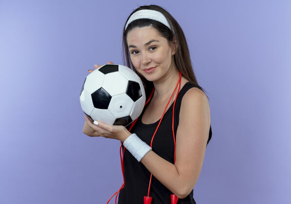 头带戴着头巾 脖子上系着跳绳 拿着足球 站在蓝色背景下微笑着看镜头的年轻健身女士球微笑健身