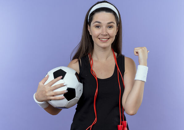 头带戴着头带 脖子上系着跳绳 拿着足球 看着镜头 微笑着握紧拳头 快乐而积极地站在蓝色背景上的年轻健身女士绳子抱着足球