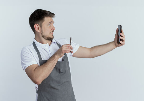 自己围裙上的理发师拿着剪刀站在白色背景上用智能手机拍照站着手机图片