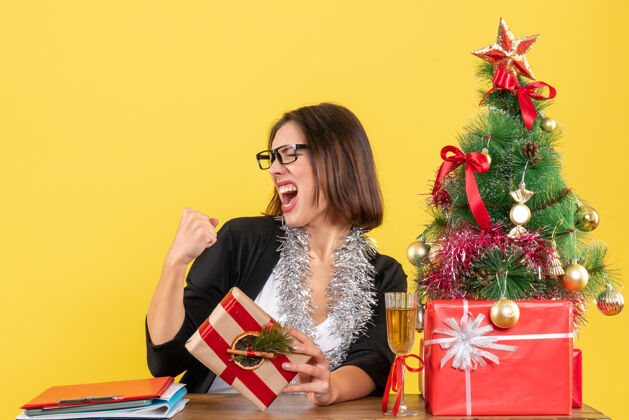 拿着一位穿着西装 戴着眼镜 骄傲地拿着礼物的美丽商务女士坐在一张桌子旁 桌子上放着一棵圣诞树快乐漂亮布置