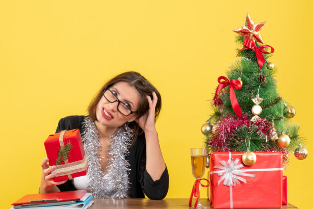 显示困惑的商务女士穿着西装 戴着眼镜展示她的礼物 坐在一张桌子旁 桌子上放着一棵圣诞树漂亮困惑眼镜