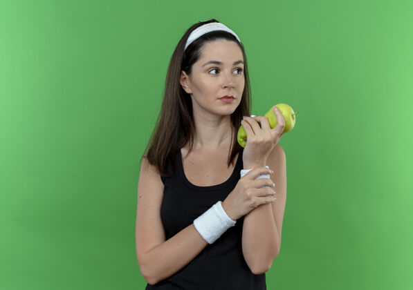 面子戴着头巾的年轻健身女士手拿青苹果 严肃的脸站在绿色的背景上 望向一边年轻苹果认真
