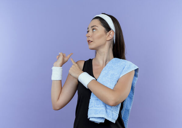 年轻戴着头巾 肩上扛着毛巾 站在蓝色背景上 指着背后看的年轻健身女士毛巾健身肩膀