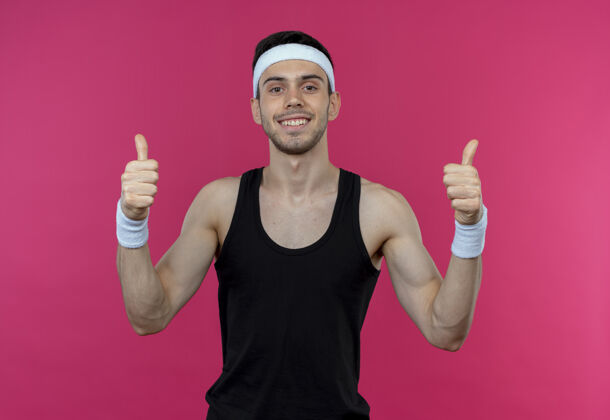 运动戴着头巾的年轻运动型男子看着镜头 微笑着 高兴而积极地竖起大拇指站在粉色背景上拇指年轻相机