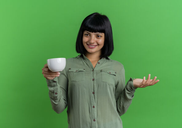绿色微笑的年轻黑发白种人女孩拿着杯子 看着在绿色背景和复制空间孤立的一面壁板举行微笑