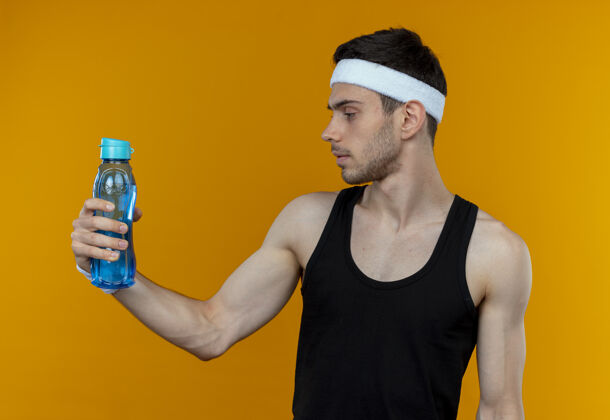站着戴着头巾的年轻人拿着一瓶水 严肃地看着它 站在橙色的背景上脸运动头带
