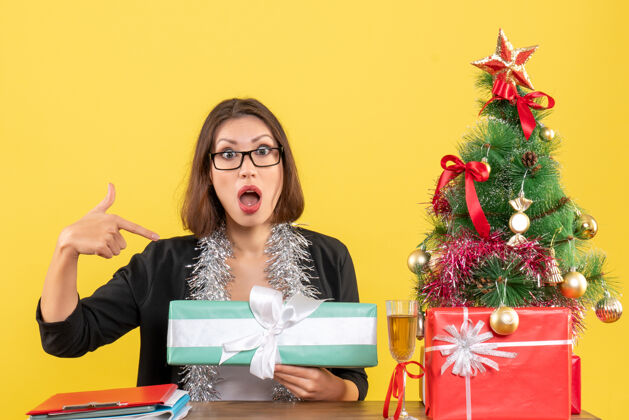 快乐一位穿着西装 戴着眼镜的商务女士惊讶地指着自己的礼物 坐在办公室的一张桌子旁 桌上放着一棵圣诞树树笔记本电脑人
