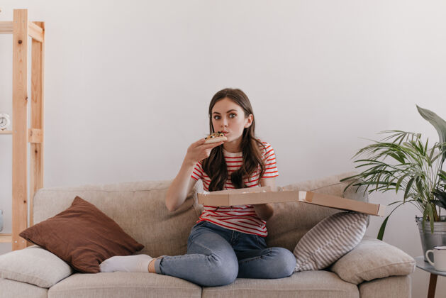 辣香肠模特坐在柔软明亮的沙发上 在温馨的家居氛围中 吃着刚刚送来的美味比萨饼年轻公寓人