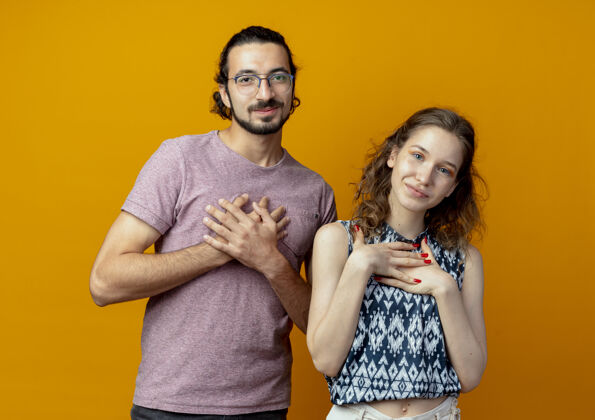 胸脯一对年轻漂亮的夫妇站在橙色的背景下 看着相机 手放在胸前 心怀感激情侣站着感情