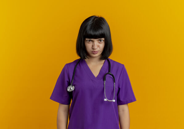 制服恼怒的年轻黑发女医生穿着制服 用听诊器看着隔离在橙色背景上的摄像头 并留有复印空间太空人恼火副本