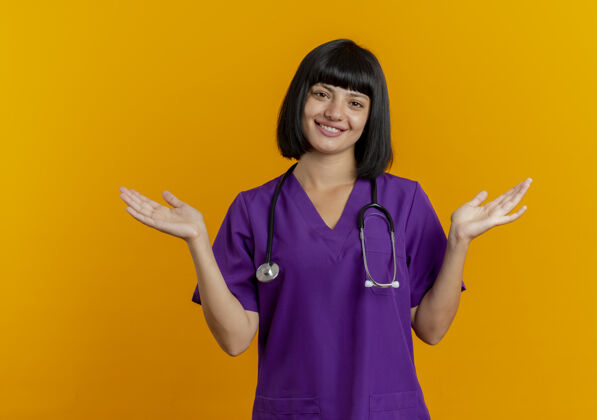 站立微笑的年轻黑发女医生穿着制服 手持听诊器站在橙色背景上 双手张开 留有复印空间微笑开放医生