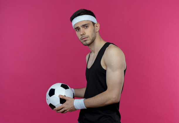相机戴着头巾 拿着足球 表情严肃地站在粉色背景下看着摄像机的年轻运动型男子球持球严肃